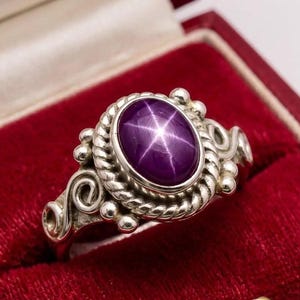 Könnte beinhalten: Silberring mit einem ovalen, tiefvioletten Edelstein, der ein sechsspitziges Sternmuster aufweist. Der Edelstein ist in eine detaillierte, seilartige Lünette gefasst. Der Ring hat dekorative Wirbel und kleine Perlenakzente.