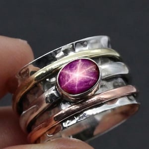 Könnte beinhalten: Ein silberner Ring mit gehämmertem Muster, mit einem zentralen, oval geformten, sternartigen Rubin-Edelstein. Der Ring hat gold- und kupferfarbene Bänder, die ein geschichtetes Design bilden. Der Ring ist ein Schmuckstück.