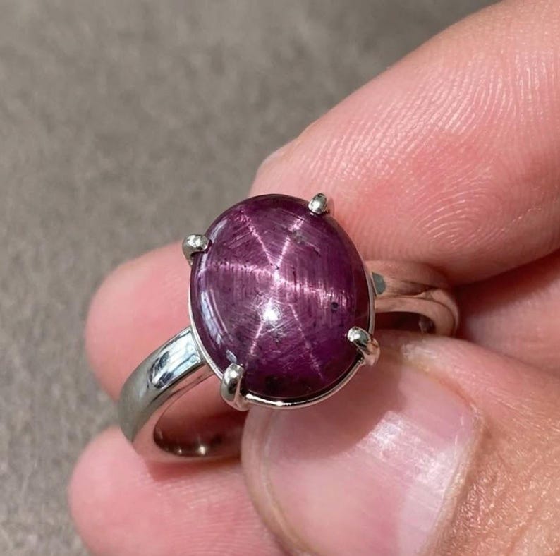 May include: A silver ring with a large, oval star ruby gemstone. The ruby is a deep, purplish-red colour, displaying a six-rayed star effect. The ring has a simple, polished silver band and four prongs securing the gemstone.