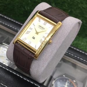 Op de afbeelding: Een goudkleurig Seiko Quartz horloge met een rechthoekige wijzerplaat en een bruine leren band. De crèmekleurige wijzerplaat heeft Romeinse cijfers en het Seiko-logo. Het horloge wordt gepresenteerd op een grijs kussen.