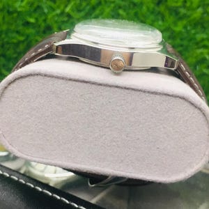 May include: A silver wristwatch with a brown leather band rests on a gray, oval-shaped cushion. The watch has a clear face and a small, round crown. The background is a blurred green.
