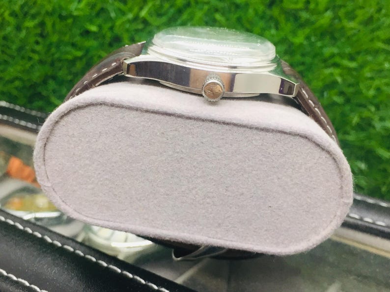 May include: A silver wristwatch with a brown leather band rests on a gray, oval-shaped cushion. The watch has a clear face and a small, round crown. The background is a blurred green.
