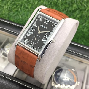 May include: A rectangular Seiko watch with a black face, silver frame, and brown leather strap. The watch face features Roman numerals and sub-dials. The watch is displayed on a grey cushion.