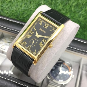 Puede incluir: Un reloj Seiko rectangular con esfera negra y detalles dorados. El reloj presenta números romanos y una correa de cuero negro. El reloj se exhibe sobre un cojín gris dentro de una caja de reloj.
