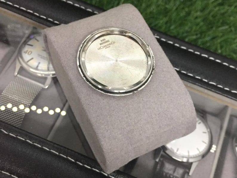 Op de afbeelding: Een zilveren horlogekast achterkant met de tekst "SWISS MADE" en "LE COULTRE & CO" wordt getoond op een grijs kussen. Andere horloges zijn zichtbaar in een zwarte horlogedoos.