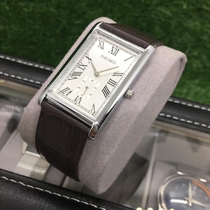 Può includere: Un orologio Seiko rettangolare con quadrante bianco, numeri romani e cinturino in pelle marrone. L'orologio ha una cassa color argento e un quadrante secondario. L'orologio è esposto su un cuscino grigio.