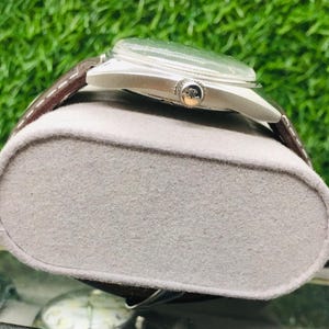 May include: A silver-toned wristwatch with a brown leather band rests on a gray, felt-covered display. The watch has a clear face and a small, decorative emblem on the side. The background is a blurred green.