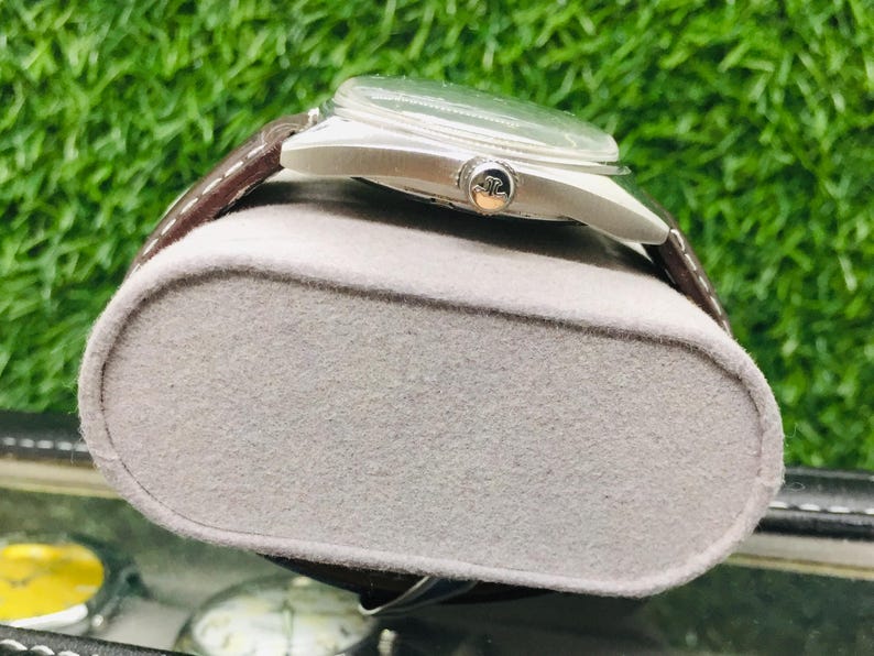 May include: A silver-toned wristwatch with a brown leather band rests on a gray, felt-covered display. The watch has a clear face and a small, decorative emblem on the side. The background is a blurred green.