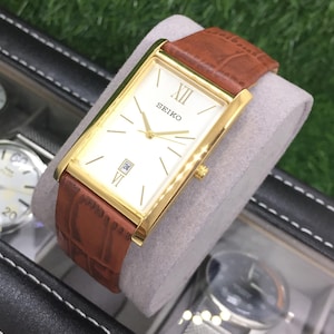 Può includere: Un orologio Seiko dorato con quadrante rettangolare e cinturino in pelle marrone. L'orologio ha un quadrante bianco con lancette e indici dorati e un datario. L'orologio è esposto su un cuscino grigio.