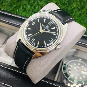 Op de afbeelding: Een zilverkleurig polshorloge met een zwarte wijzerplaat en een zwarte leren band. Het horloge heeft witte uurmarkeringen en wijzers, met de merknaam "Jaeger-LeCoultre". Het horloge wordt gepresenteerd in een zwarte horlogedoos.