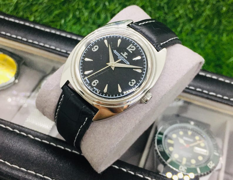 Op de afbeelding: Een zilverkleurig polshorloge met een zwarte wijzerplaat en een zwarte leren band. Het horloge heeft witte uurmarkeringen en wijzers, met de merknaam "Jaeger-LeCoultre". Het horloge wordt gepresenteerd in een zwarte horlogedoos.
