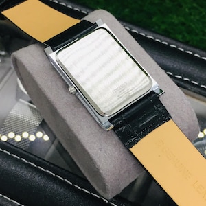 K&ouml;nnte beinhalten: Eine rechteckige silberfarbene Uhr mit schwarzem Lederarmband und hellbrauner Unterseite. Die Uhr liegt auf einem grauen Kissen. Das Zifferblatt ist nicht sichtbar. Die Uhr befindet sich in einem schwarzen Etui.