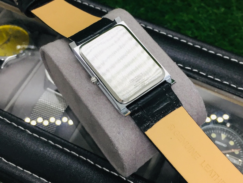 K&ouml;nnte beinhalten: Eine rechteckige silberfarbene Uhr mit schwarzem Lederarmband und hellbrauner Unterseite. Die Uhr liegt auf einem grauen Kissen. Das Zifferblatt ist nicht sichtbar. Die Uhr befindet sich in einem schwarzen Etui.