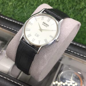 May include: A silver Seiko Quartz Slim watch with a white face and black leather band. The watch features Roman numeral hour markers and a second hand. The watch is displayed on a gray cushion inside a black case.