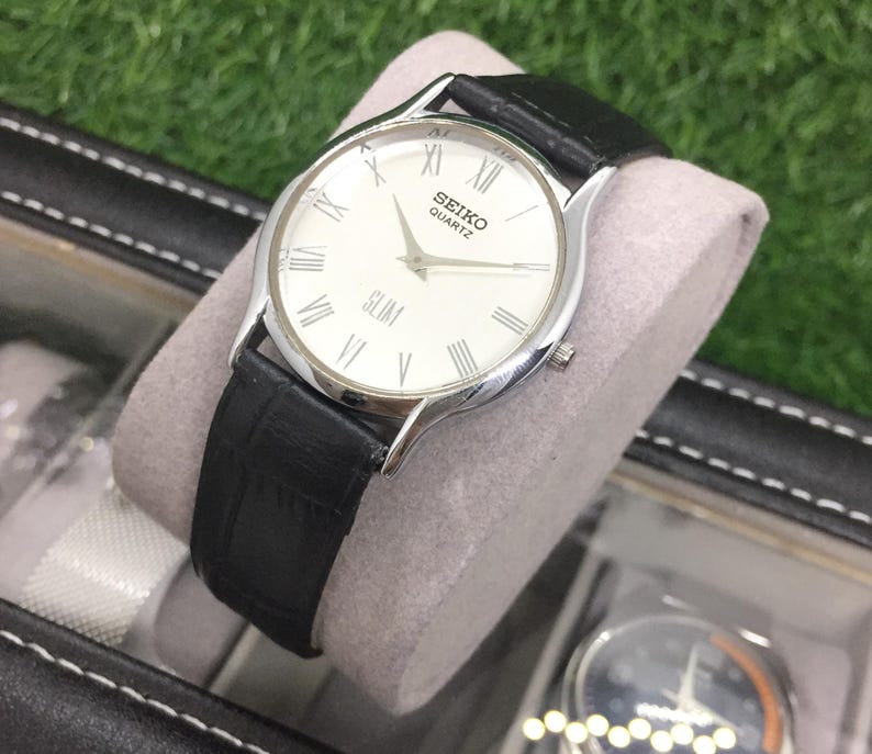 May include: A silver Seiko Quartz Slim watch with a white face and black leather band. The watch features Roman numeral hour markers and a second hand. The watch is displayed on a gray cushion inside a black case.