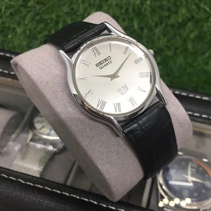 May include: A silver Seiko Quartz Slim watch with a white face and black leather band. The watch features Roman numeral hour markers and a second hand. The watch is displayed on a gray cushion.