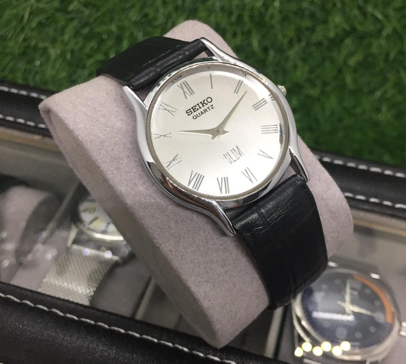 May include: A silver Seiko Quartz Slim watch with a white face and black leather band. The watch features Roman numeral hour markers and a second hand. The watch is displayed on a gray cushion.