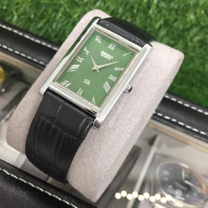 May include: A rectangular Seiko Quartz watch with a green face and silver-tone case. The watch features Roman numeral hour markers and a black leather band. The watch is displayed on a gray cushion.