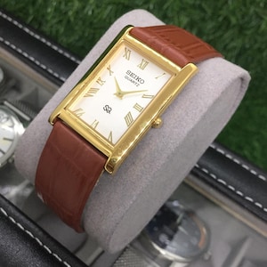 Puede incluir: Reloj Seiko Quartz dorado con esfera rectangular y correa de cuero marrón. La esfera es blanca con agujas doradas y números romanos. La correa es de color marrón intenso.