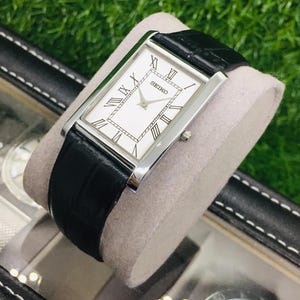 K&ouml;nnte beinhalten: Eine rechteckige Seiko-Uhr mit wei&szlig;em Zifferblatt, schwarzen r&ouml;mischen Ziffern und einem schwarzen Lederarmband. Die Uhr hat ein silberfarbenes Geh&auml;use und wird auf einem hellgrauen Kissen pr&auml;sentiert. Die Uhr befindet sich in einer schwarzen Box.