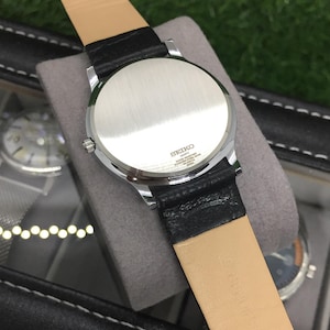May include: A silver-toned Seiko quartz watch with a black leather strap and a light brown underside. The watch is displayed on a gray surface, with the back of the watch facing the camera. The watch case is round.