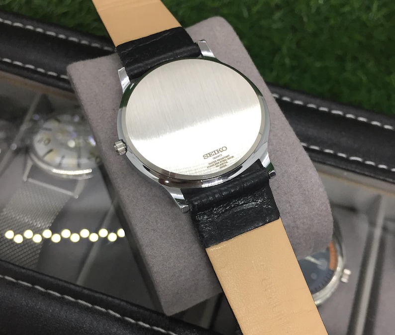 May include: A silver-toned Seiko quartz watch with a black leather strap and a light brown underside. The watch is displayed on a gray surface, with the back of the watch facing the camera. The watch case is round.
