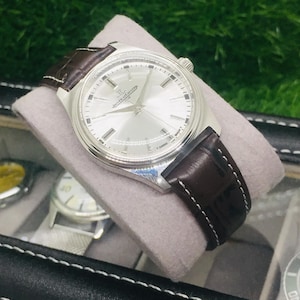 May include: A silver-toned wristwatch with a light-colored face and dark brown leather band. The watch is displayed on a gray cushion inside a black watch case. The watch face has silver hands and markers.