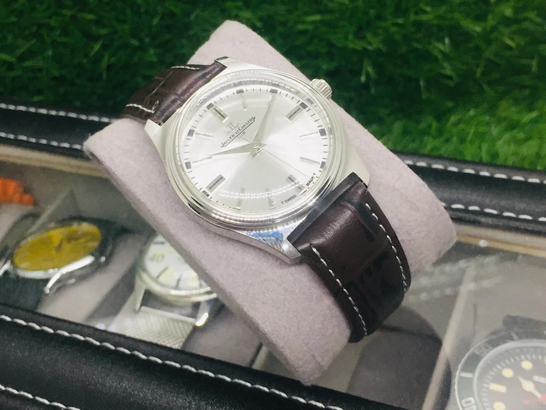 May include: A silver-toned wristwatch with a light-colored face and dark brown leather band. The watch is displayed on a gray cushion inside a black watch case. The watch face has silver hands and markers.