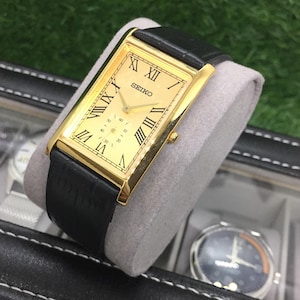 Peut inclure: Une montre Seiko dorée avec un cadran rectangulaire et un bracelet en cuir noir. Le cadran de la montre présente des chiffres romains et un sous-cadran. La montre est présentée sur un coussin gris dans un étui noir.