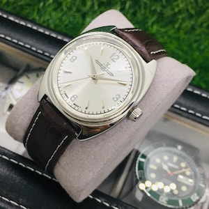 May include: A silver-toned wristwatch with a white face and brown leather band. The watch features a round face with black numerals and hands. The band is dark brown with white stitching. The watch is displayed on a gray pillow inside a black case.