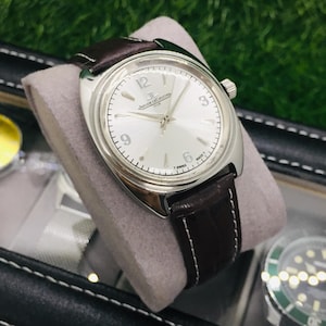 May include: A silver-toned wristwatch with a light-colored face and dark brown leather band. The watch features a round face with black numerals and hands. The band is stitched with white thread. The watch is displayed on a gray cushion.