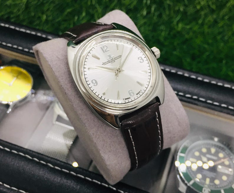 May include: A silver-toned wristwatch with a light-colored face and dark brown leather band. The watch features a round face with black numerals and hands. The band is stitched with white thread. The watch is displayed on a gray cushion.