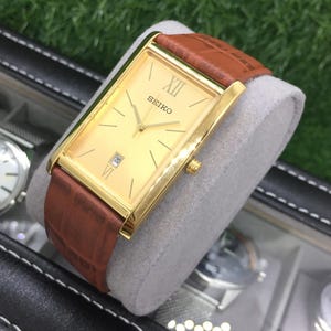May include: A gold-toned Seiko watch with a rectangular face and brown leather band. The watch face displays Roman numerals and a date window. The watch is displayed on a gray cushion.