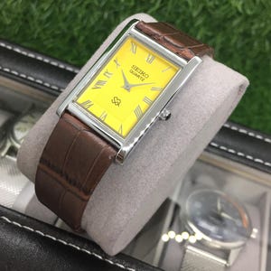 Puede incluir: Un reloj Seiko Quartz rectangular con esfera amarilla brillante y correa de cuero marrón. El reloj tiene bordes plateados y números romanos. El reloj se muestra sobre un cojín gris.