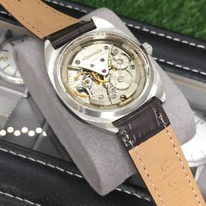 May include: A vintage wristwatch with a silver case and a brown leather strap. The watch's intricate internal mechanism is visible through the clear back. The strap is marked with the word "Nakata".