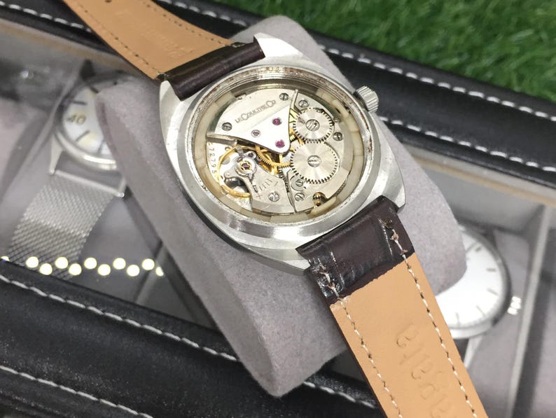 May include: A vintage wristwatch with a silver case and a brown leather strap. The watch's intricate internal mechanism is visible through the clear back. The strap is marked with the word "Nakata".