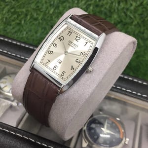 Puede incluir: Un reloj Seiko Quartz rectangular con esfera plateada y correa de cuero marrón. El reloj tiene una caja plateada y una correa de cuero marrón texturizado. El reloj se muestra sobre un cojín gris.