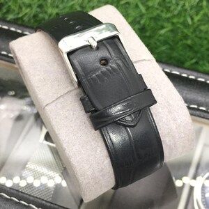 May include: A black leather watch strap with a silver buckle is displayed on a gray cushion. The strap has a textured surface and a small loop to secure the excess length. The watch strap is part of a watch collection.
