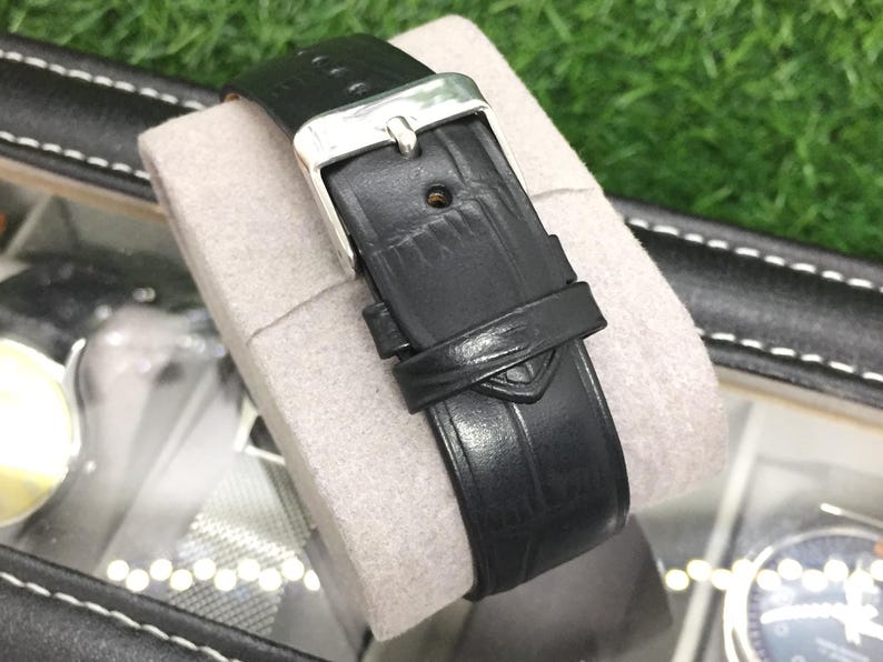 May include: A black leather watch strap with a silver buckle is displayed on a gray cushion. The strap has a textured surface and a small loop to secure the excess length. The watch strap is part of a watch collection.