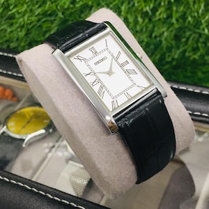 K&ouml;nnte beinhalten: Eine rechteckige Seiko-Uhr mit wei&szlig;em Zifferblatt, schwarzen r&ouml;mischen Ziffern und einem schwarzen Lederarmband. Die Uhr wird auf einem grauen Kissen in einem schwarzen Uhrenetui pr&auml;sentiert. Das Zifferblatt tr&auml;gt die Marke "SEIKO".