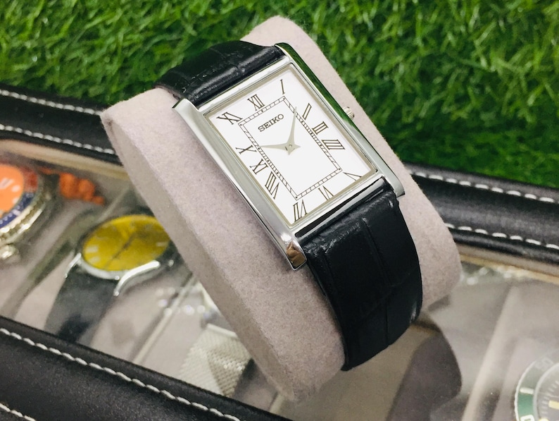 K&ouml;nnte beinhalten: Eine rechteckige Seiko-Uhr mit wei&szlig;em Zifferblatt, schwarzen r&ouml;mischen Ziffern und einem schwarzen Lederarmband. Die Uhr wird auf einem grauen Kissen in einem schwarzen Uhrenetui pr&auml;sentiert. Das Zifferblatt tr&auml;gt die Marke "SEIKO".