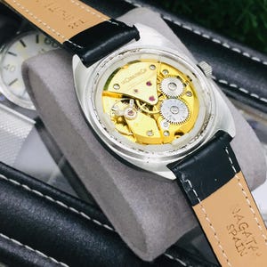 Op de afbeelding: Close-up van een zilverkleurig polshorloge met een zwarte leren band. De achterkant van het horloge is zichtbaar en toont de ingewikkelde goudkleurige interne mechanismen. De band is gemarkeerd met "NAGATA SPAIN".