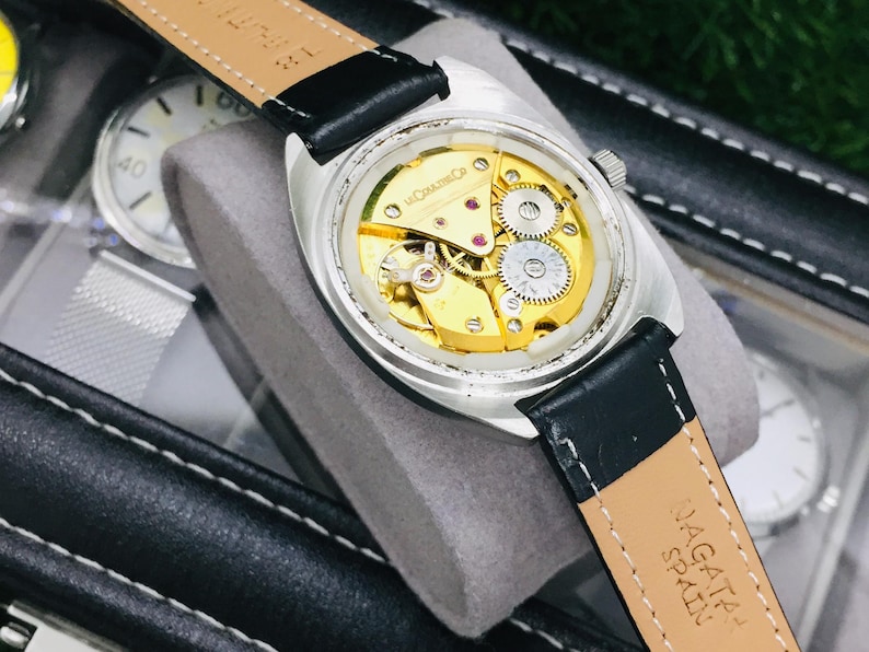Op de afbeelding: Close-up van een zilverkleurig polshorloge met een zwarte leren band. De achterkant van het horloge is zichtbaar en toont de ingewikkelde goudkleurige interne mechanismen. De band is gemarkeerd met "NAGATA SPAIN".