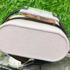K&ouml;nnte beinhalten: Eine rechteckige Armbanduhr mit silberfarbenem Geh&auml;use und schwarzem Lederarmband. Die Uhr wird auf einem grauen, oval geformten Uhrenhalter pr&auml;sentiert. Der Hintergrund zeigt gr&uuml;nes Gras.