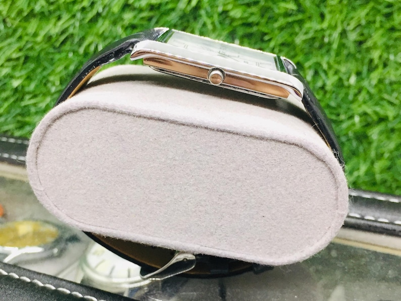 K&ouml;nnte beinhalten: Eine rechteckige Armbanduhr mit silberfarbenem Geh&auml;use und schwarzem Lederarmband. Die Uhr wird auf einem grauen, oval geformten Uhrenhalter pr&auml;sentiert. Der Hintergrund zeigt gr&uuml;nes Gras.
