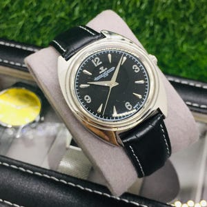 Op de afbeelding: Een zilverkleurig horloge met een zwarte wijzerplaat en een zwarte leren band. De wijzerplaat heeft witte uurmarkeringen en wijzers, met de merknaam bovenaan. Het horloge wordt getoond op een grijs kussen.