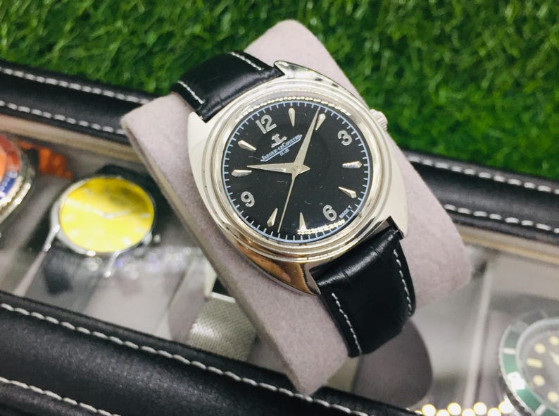 Op de afbeelding: Een zilverkleurig horloge met een zwarte wijzerplaat en een zwarte leren band. De wijzerplaat heeft witte uurmarkeringen en wijzers, met de merknaam bovenaan. Het horloge wordt getoond op een grijs kussen.