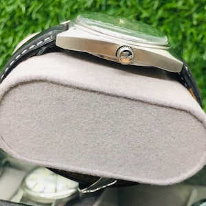 Op de afbeelding: Een zilverkleurig polshorloge met een groene wijzerplaat en een zwarte leren band rust op een grijze vilten display. Het horloge heeft een ronde wijzerplaat en een zilveren kroon. De achtergrond is een vage groene kleur.