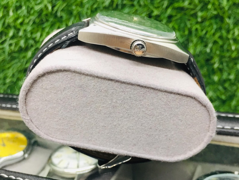 Op de afbeelding: Een zilverkleurig polshorloge met een groene wijzerplaat en een zwarte leren band rust op een grijze vilten display. Het horloge heeft een ronde wijzerplaat en een zilveren kroon. De achtergrond is een vage groene kleur.