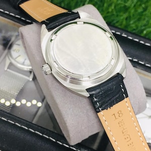 Op de afbeelding: Close-up van een zilverkleurig polshorloge met een zwarte leren band. Het horloge staat tegen een grijze achtergrond, met andere horloges zichtbaar op de achtergrond. De band is gemarkeerd met "GENUINE LEATHER" en het nummer "18".